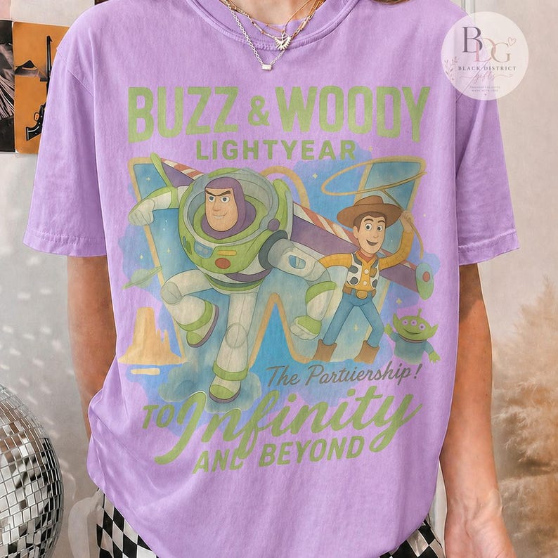 May include: A light purple t-shirt featuring Buzz Lightyear and Woody from Toy Story. The graphic includes the text "Buzz & Woody Lightyear" and "To Infinity and Beyond". The shirt has short sleeves.