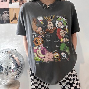 May include: Dark gray t-shirt featuring a colorful graphic of characters from the animated movie Toy Story. The design includes Woody, Buzz Lightyear, Jessie, and other characters with their names written below. The shirt has a crew neck and short sleeves.