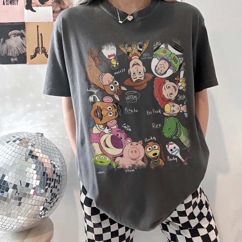 May include: Dark gray t-shirt featuring a colorful graphic of characters from the animated movie Toy Story. The design includes Woody, Buzz Lightyear, Jessie, and other characters with their names written below. The shirt has a crew neck and short sleeves.