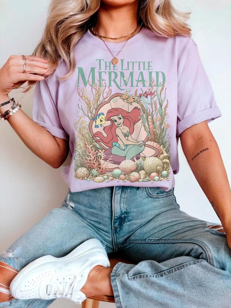 Op de afbeelding: Een lila T-shirt met een afbeelding van "De Kleine Zeemeermin". Het ontwerp bevat Ariel, Bot en andere zeedieren in een schelp. De tekst "The Little Mermaid" staat in het groen boven de illustratie.