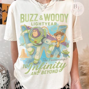 May include: Cream-colored t-shirt featuring a graphic of Buzz Lightyear and Woody from Toy Story. The design includes the text "BUZZ & WOODY LIGHTYEAR" and "TO INFINITY AND BEYOND" in green and blue hues. A small green alien is also featured.