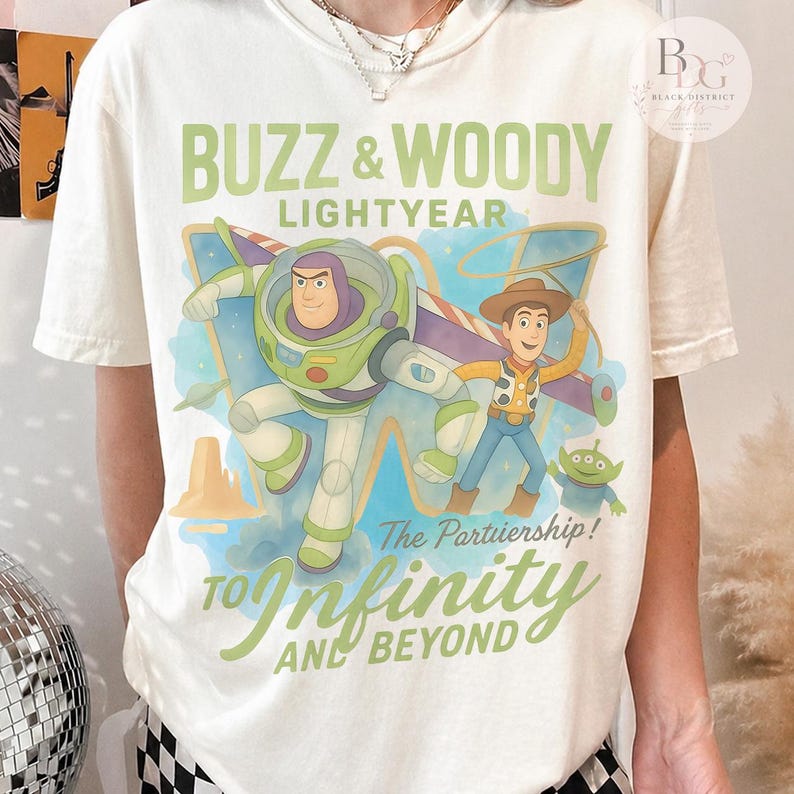 May include: Cream-colored t-shirt featuring a graphic of Buzz Lightyear and Woody from Toy Story. The design includes the text "BUZZ & WOODY LIGHTYEAR" and "TO INFINITY AND BEYOND" in green and blue hues. A small green alien is also featured.