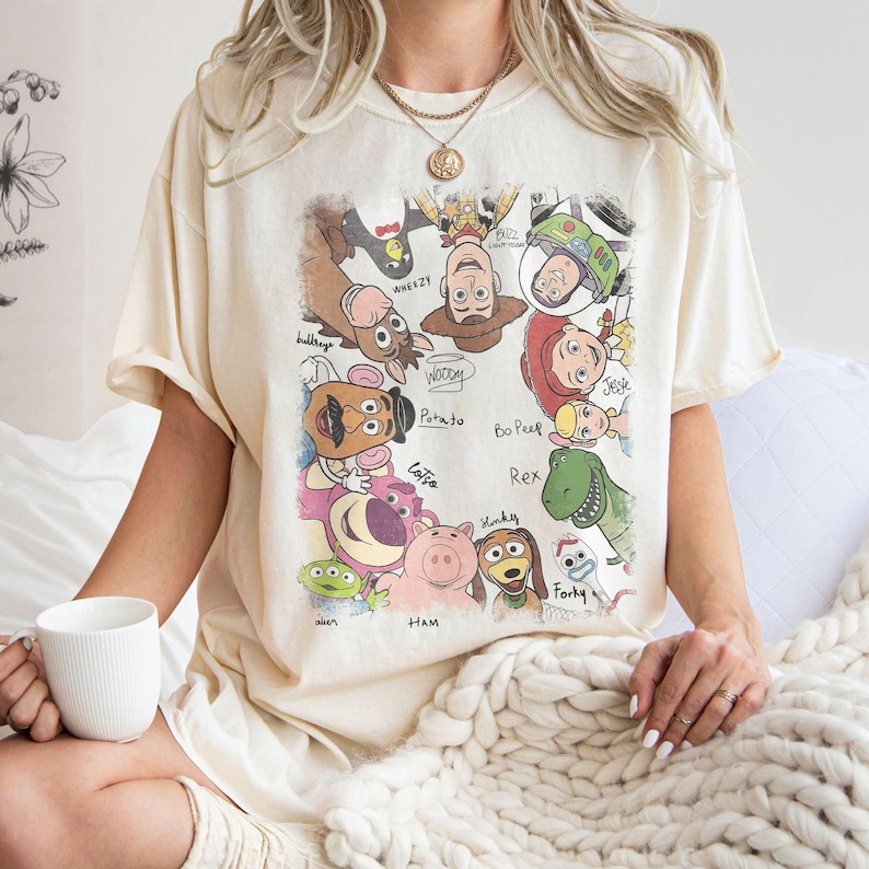 May include: Cream-colored t-shirt featuring a colorful graphic of various Toy Story characters, including Woody, Buzz Lightyear, and Rex. The design includes character names in a handwritten style. The shirt has short sleeves and a relaxed fit.
