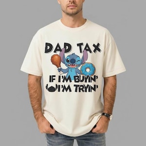 May include: Cream-colored t-shirt featuring a cartoon character holding a drumstick and a donut. The shirt has the text "DAD TAX" above the character and "IF I'M BUYIN' I'M TRYIN'" below. The character is blue with large ears.