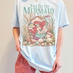 Op de afbeelding: Lichtblauw T-shirt met een afbeelding van Ariel uit De Kleine Zeemeermin, omringd door schelpen en waterplanten. De tekst "The Little Mermaid" staat boven de illustratie. Het shirt wordt gecombineerd met een rood-witte geruite short.