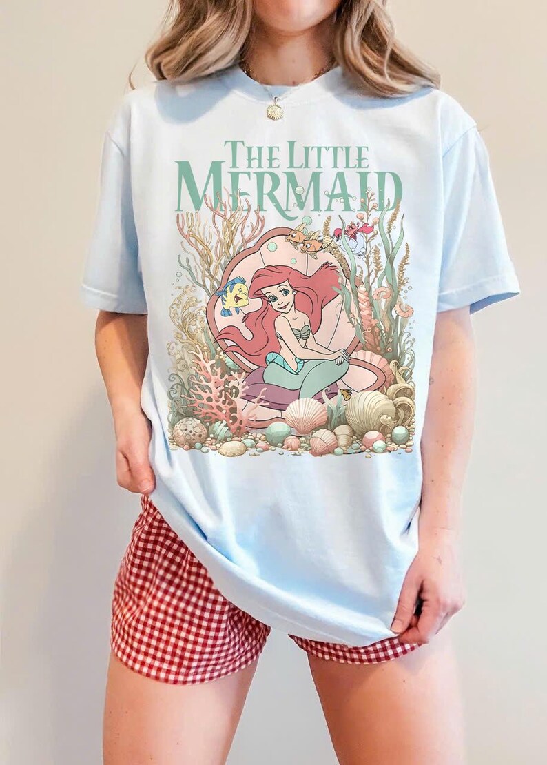 Op de afbeelding: Lichtblauw T-shirt met een afbeelding van Ariel uit De Kleine Zeemeermin, omringd door schelpen en waterplanten. De tekst "The Little Mermaid" staat boven de illustratie. Het shirt wordt gecombineerd met een rood-witte geruite short.