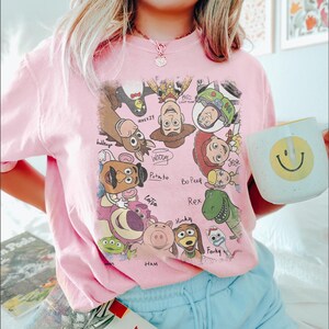 May include: A light pink t-shirt featuring a colorful graphic of characters from the animated movie Toy Story. The shirt also has a white mug with a yellow smiley face. The shirt is paired with light blue shorts.