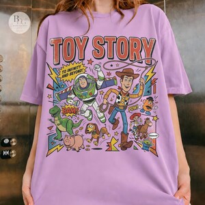May include: A lilac-colored t-shirt featuring a colorful graphic from the animated film "Toy Story." The design includes the text "TOY STORY" and various characters like Buzz Lightyear and Woody. The phrase "TO INFINITY AND BEYOND!" is also present.