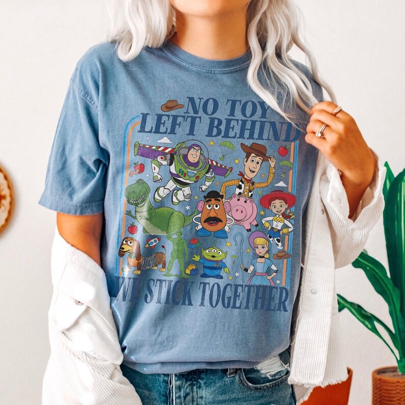 May include: A blue t-shirt featuring a colorful graphic of characters from the animated film "Toy Story." The design includes the text "NO TOY LEFT BEHIND" and "WE STICK TOGETHER." The shirt is a light blue color.