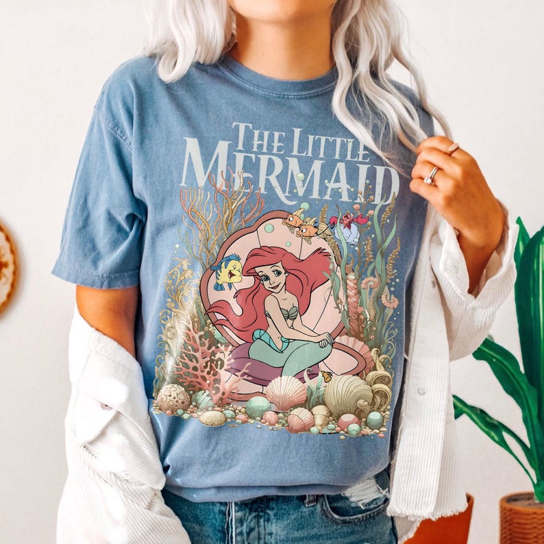 Op de afbeelding: Een lichtblauw T-shirt met de tekst "The Little Mermaid" en een illustratie van Ariel, Botje en Sebastiaan. De afbeelding bevat koraal, schelpen en andere zeedieren. Het T-shirt is lichtblauw van kleur.