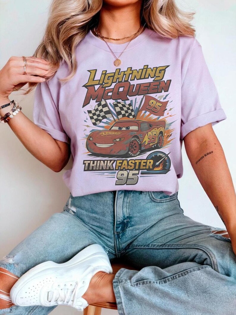 K&ouml;nnte beinhalten: Ein helllila T-Shirt mit einer Grafik von Lightning McQueen aus dem Film Cars. Das Design enth&auml;lt den Text "Lightning McQueen", "95" und "Think Faster". Das Shirt wird mit einer blauen Jeans getragen.
