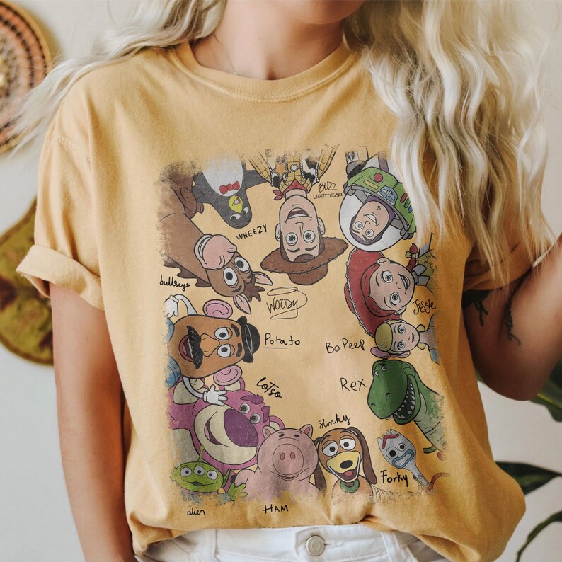 May include: Mustard yellow t-shirt featuring a graphic of various Toy Story characters, including Woody, Buzz Lightyear, and Jessie. The names of the characters are written below them. The shirt has short sleeves and a crew neck.