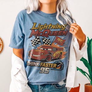 K&ouml;nnte beinhalten: Hellblaues T-Shirt mit Lightning McQueen aus dem Film Cars. Die Grafik enth&auml;lt den Text "Lightning McQueen", "Think Faster" und die Nummer "95". Das Design zeigt ein rotes Rennauto mit Zielflaggen und Flammen.