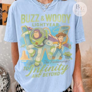 May include: Light blue t-shirt featuring Buzz Lightyear and Woody from Toy Story. The graphic includes the text "Buzz & Woody Lightyear" and "To Infinity and Beyond". The design has a cartoon style with a blue and green color scheme.