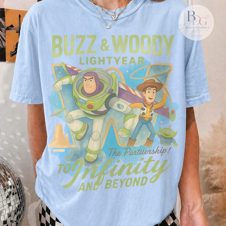 May include: Light blue t-shirt featuring Buzz Lightyear and Woody from Toy Story. The graphic includes the text "Buzz & Woody Lightyear" and "To Infinity and Beyond". The design has a cartoon style with a blue and green color scheme.