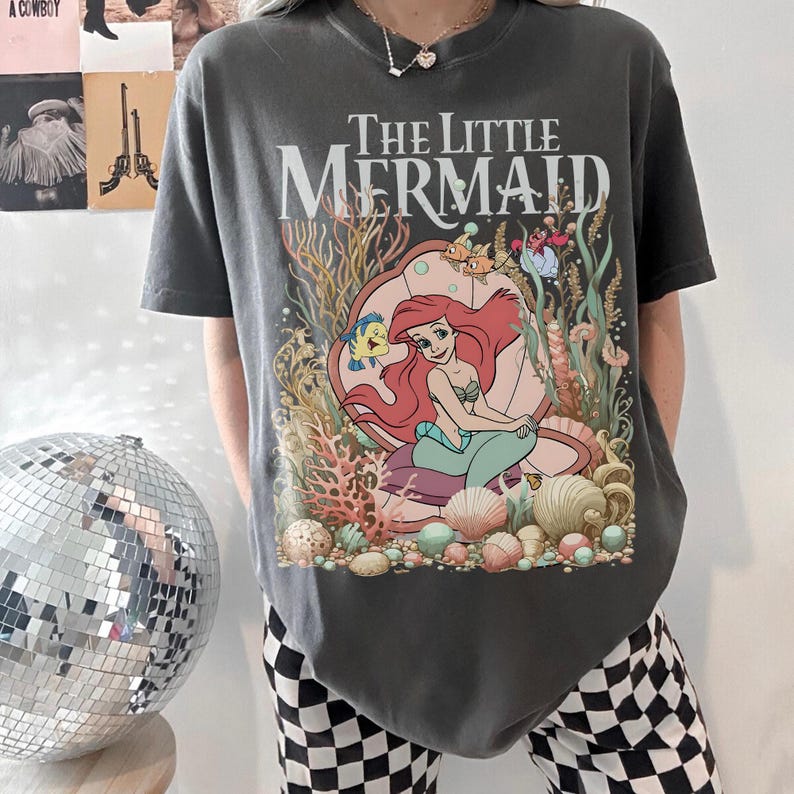 Op de afbeelding: Donkergrijs T-shirt met de tekst "The Little Mermaid" boven een kleurrijke afbeelding van Ariel in een schelp, omringd door koraal, vissen en schelpen. Het shirt heeft korte mouwen en een relaxte pasvorm.