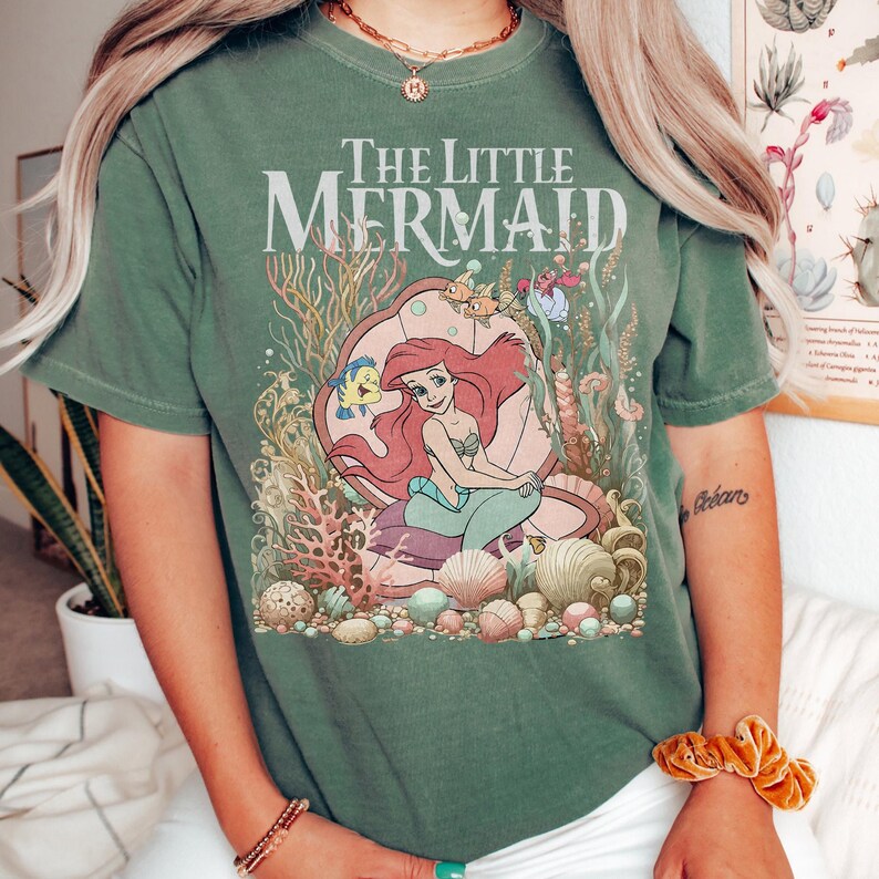 Op de afbeelding: Groen T-shirt met een afbeelding van Disney's De Kleine Zeemeermin. Het ontwerp bevat Ariel, Botje en andere zeedieren. De tekst "The Little Mermaid" staat boven de illustratie gedrukt. Het shirt heeft een relaxte pasvorm.