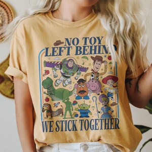 May include: Mustard yellow t-shirt featuring a colorful graphic of animated characters with the text "NO TOY LEFT BEHIND WE STICK TOGETHER". The design includes a variety of cartoon figures in a retro style.