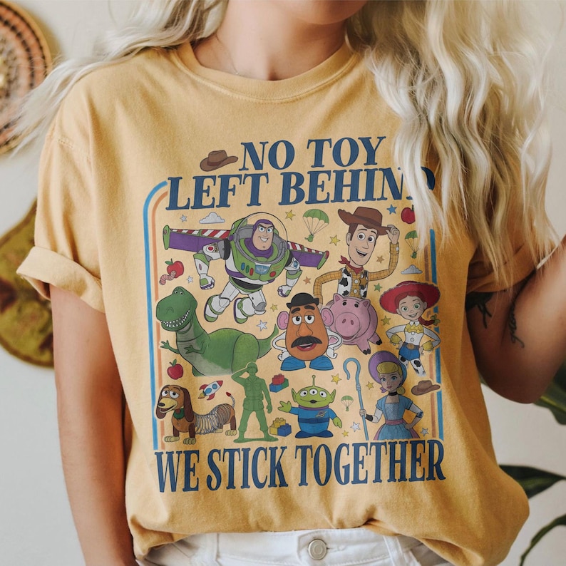 May include: Mustard yellow t-shirt featuring a colorful graphic of animated characters with the text "NO TOY LEFT BEHIND WE STICK TOGETHER". The design includes a variety of cartoon figures in a retro style.