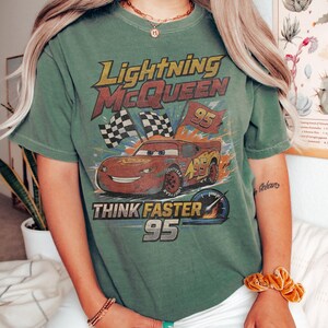 K&ouml;nnte beinhalten: Gr&uuml;nes T-Shirt mit Lightning McQueen-Design aus dem Film Cars. Das Design zeigt den Text "Lightning McQueen", "Think Faster" und die Nummer "95", sowie Zielflaggen und einen Tachometer.
