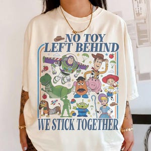 May include: Cream-colored t-shirt featuring a colorful graphic of characters from the animated film Toy Story. The text "NO TOY LEFT BEHIND WE STICK TOGETHER" is printed above and below the characters in a blue font.