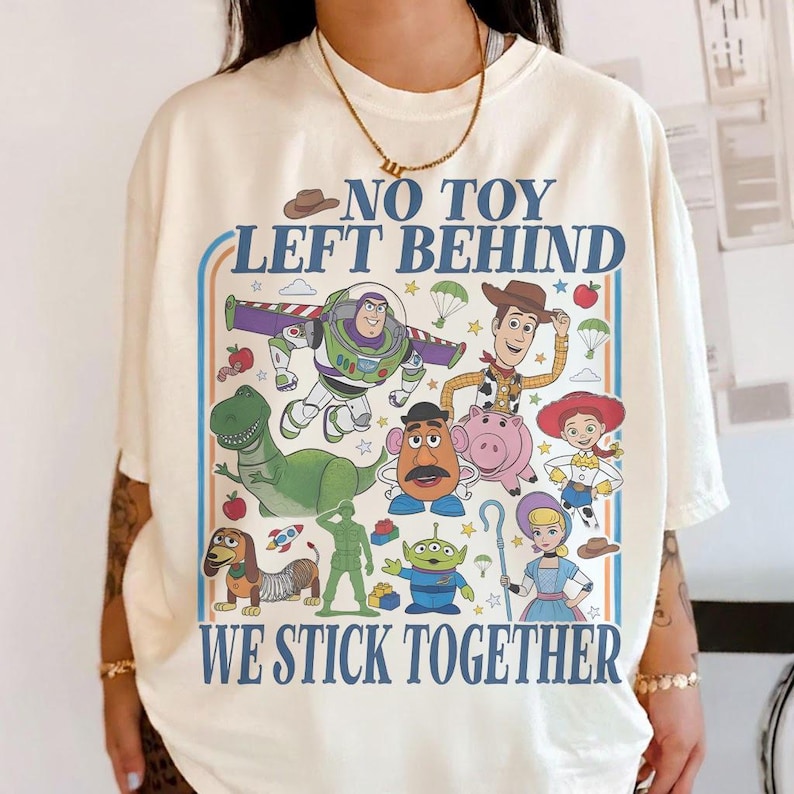 May include: Cream-colored t-shirt featuring a colorful graphic of characters from the animated film Toy Story. The text "NO TOY LEFT BEHIND WE STICK TOGETHER" is printed above and below the characters in a blue font.