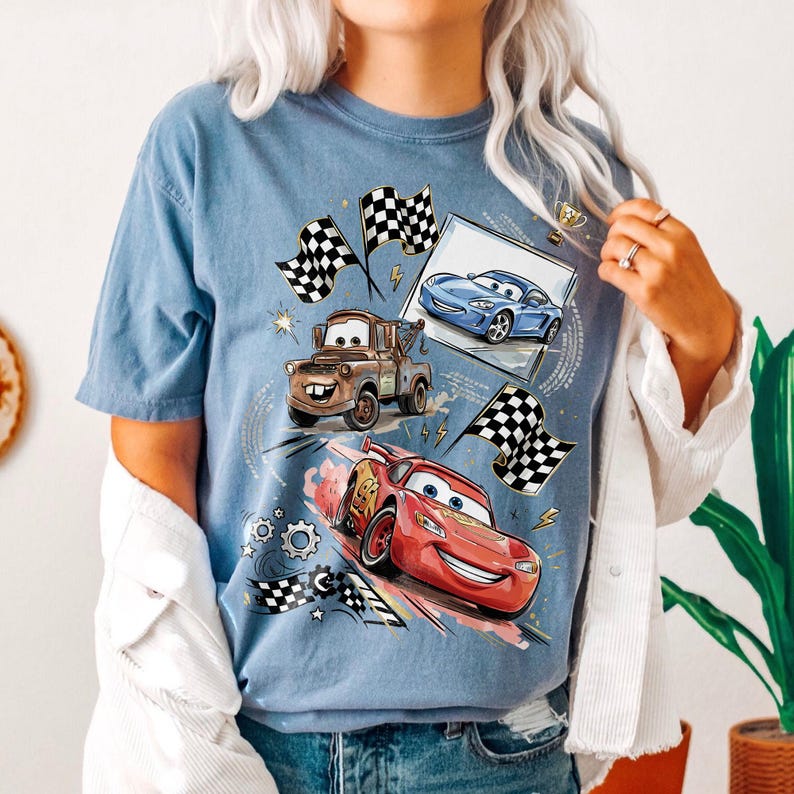 Op de afbeelding: Lichtblauw T-shirt met cartoonauto's, waaronder Bliksem McQueen, Takel en een blauwe raceauto. Het ontwerp bevat geblokte vlaggen, een trofee en race-elementen. Het shirt is lichtblauw.