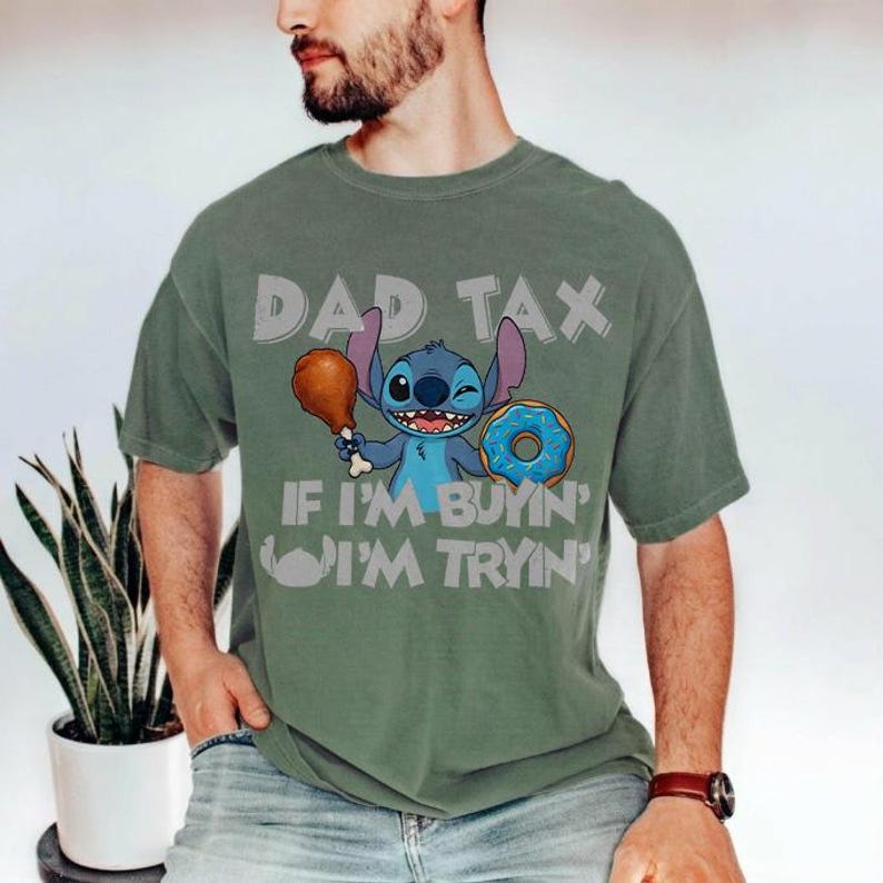 May include: Olive green t-shirt featuring a cartoon character holding a drumstick and a donut. The text "DAD TAX" and "IF I'M BUYIN' I'M TRYIN'" are printed above and below the character. The shirt is a casual style.