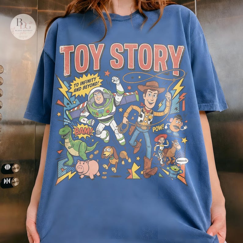 May include: Blue t-shirt featuring a colorful graphic of characters from the animated film "Toy Story." The design includes Buzz Lightyear, Woody, and other characters with the text "TOY STORY" and "TO INFINITY AND BEYOND!"