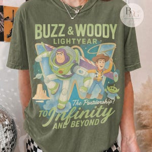 May include: Olive green t-shirt featuring Buzz Lightyear and Woody from Toy Story. The graphic includes the text "BUZZ & WOODY LIGHTYEAR" and "TO Infinity AND BEYOND". The design showcases the characters in a colorful, cartoon style.