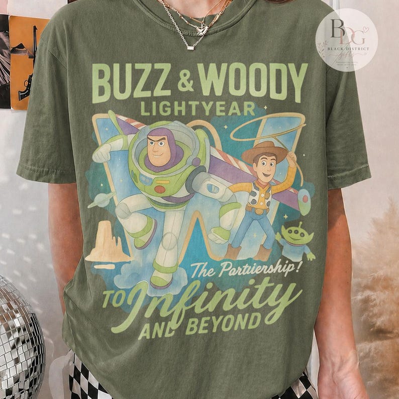 May include: Olive green t-shirt featuring Buzz Lightyear and Woody from Toy Story. The graphic includes the text "BUZZ & WOODY LIGHTYEAR" and "TO Infinity AND BEYOND". The design showcases the characters in a colorful, cartoon style.