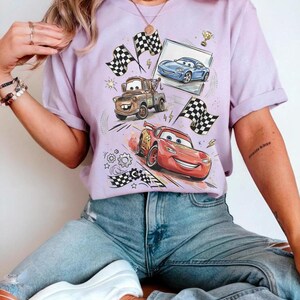 Op de afbeelding: Een lichtpaars t-shirt met een afbeelding van geanimeerde raceauto's, waaronder Bliksem McQueen, Takel en een blauwe auto. Het ontwerp bevat geblokte vlaggen, een trofee en een ingelijste afbeelding van een auto. Het shirt is gemaakt van zacht materiaal.