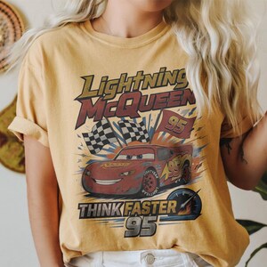 K&ouml;nnte beinhalten: Senfgelbes T-Shirt mit einer Grafik von Lightning McQueen aus dem Film Cars. Das Design enth&auml;lt den Text "Lightning McQueen", "Think Faster" und die Nummer "95" sowie karierte Flaggen.