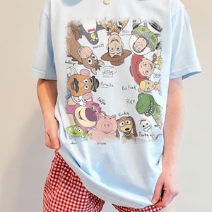 May include: Light blue t-shirt featuring a colorful graphic of Toy Story characters, including Woody, Buzz Lightyear, and Jessie. The shirt has short sleeves and a crew neck. The person is wearing red and white checkered shorts.