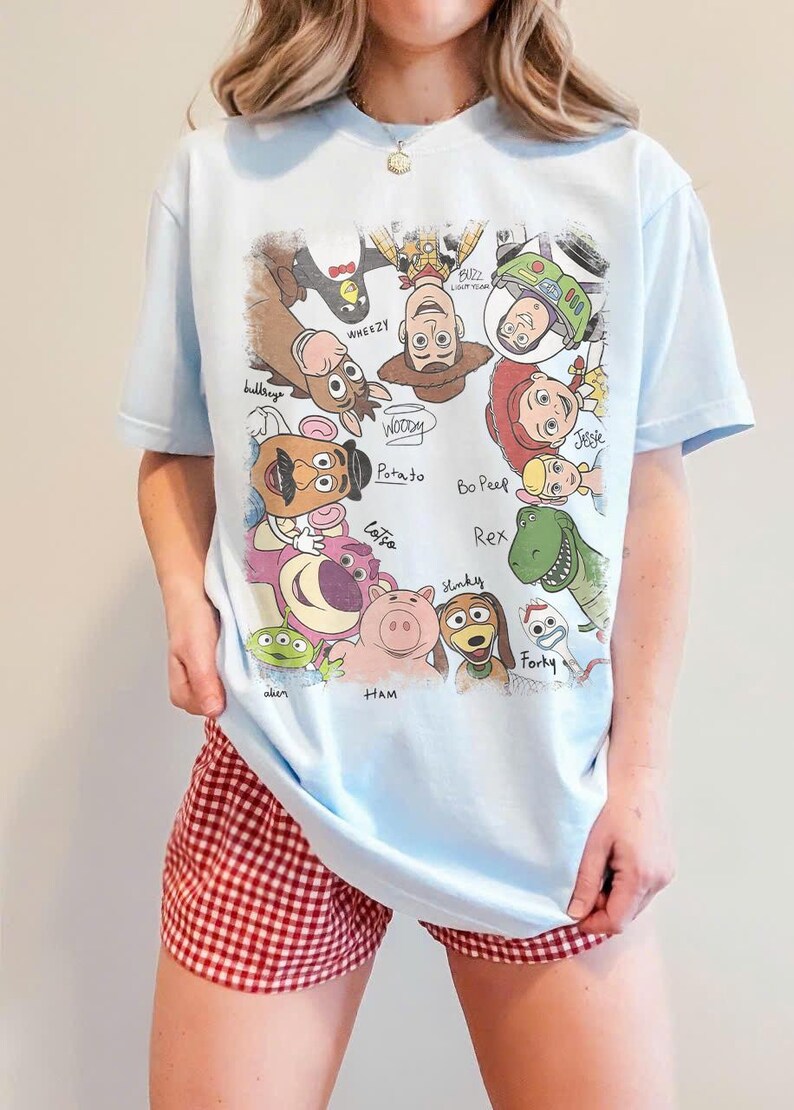 May include: Light blue t-shirt featuring a colorful graphic of Toy Story characters, including Woody, Buzz Lightyear, and Jessie. The shirt has short sleeves and a crew neck. The person is wearing red and white checkered shorts.