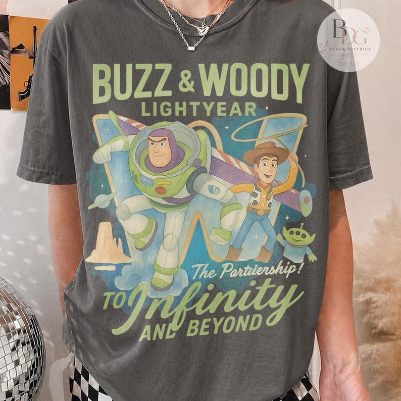 May include: Dark gray t-shirt featuring Buzz Lightyear and Woody from Toy Story. The graphic includes the text "BUZZ & WOODY LIGHTYEAR" and "TO Infinity AND BEYOND". The design showcases the characters in a colorful, cartoon style.