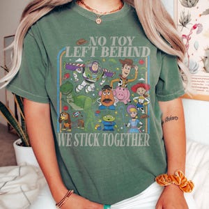May include: Green t-shirt featuring a colorful graphic of characters from the animated film Toy Story. The design includes Buzz Lightyear, Woody, and other characters, with the text "NO TOY LEFT BEHIND WE STICK TOGETHER".
