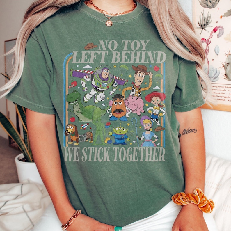 May include: Green t-shirt featuring a colorful graphic of characters from the animated film Toy Story. The design includes Buzz Lightyear, Woody, and other characters, with the text "NO TOY LEFT BEHIND WE STICK TOGETHER".