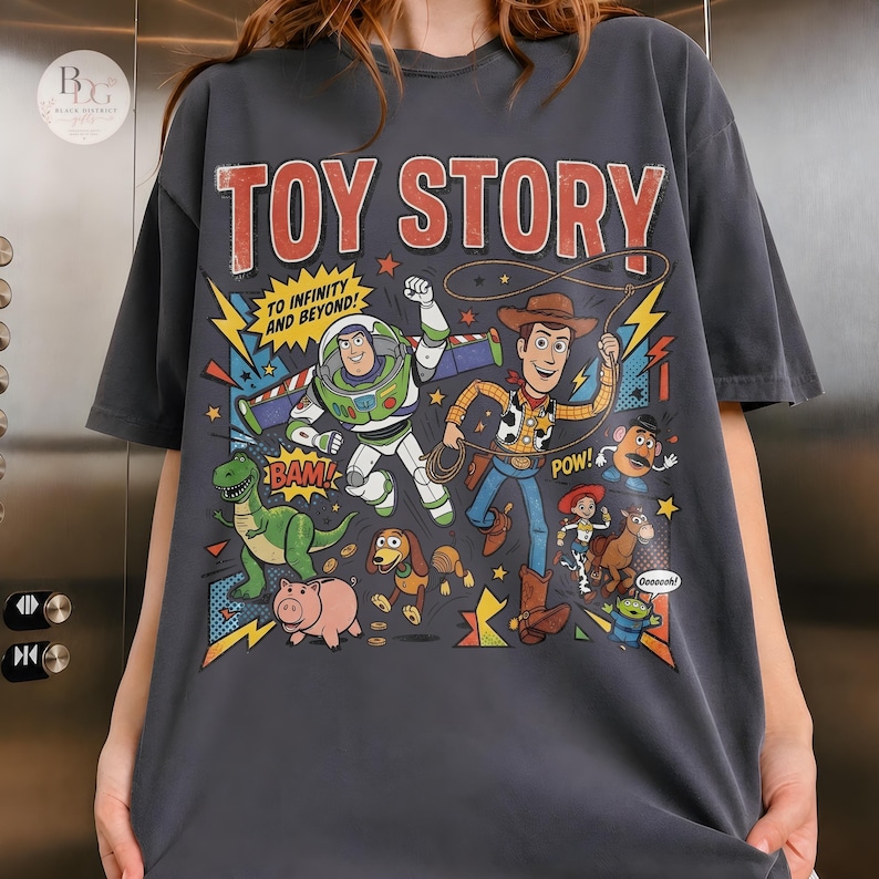 May include: Dark gray t-shirt featuring a colorful graphic of characters from the animated film "Toy Story." The design includes the words "TOY STORY" in red, and "TO INFINITY AND BEYOND!" in yellow. The shirt showcases Woody, Buzz Lightyear, and other characters.