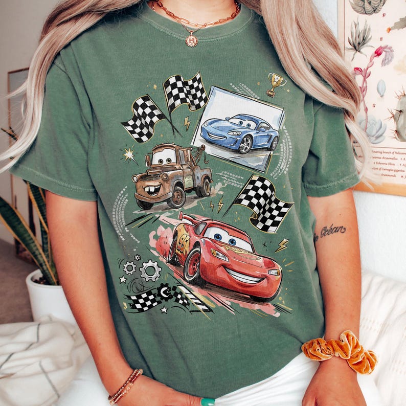 Op de afbeelding: Groen t-shirt met een afbeelding van geanimeerde raceauto's, waaronder Lightning McQueen, Mater en een blauwe auto, met geblokte vlaggen en een trofee. Het ontwerp is gecentreerd op het shirt, met een vintage, licht vervaagde look.