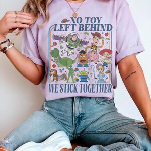 May include: A lilac-colored t-shirt featuring a colorful graphic of characters from the animated film "Toy Story." The design includes Buzz Lightyear, Woody, and other characters, with the text "NO TOY LEFT BEHIND" and "WE STICK TOGETHER."