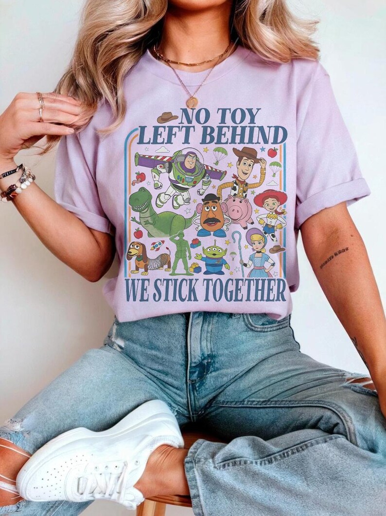 May include: A lilac-colored t-shirt featuring a colorful graphic of characters from the animated film "Toy Story." The design includes Buzz Lightyear, Woody, and other characters, with the text "NO TOY LEFT BEHIND" and "WE STICK TOGETHER."