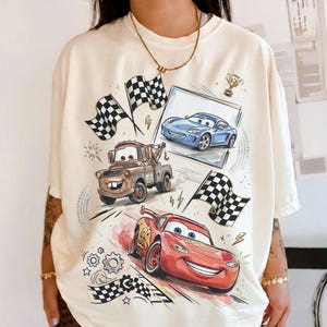Op de afbeelding: Cr&egrave;mekleurig T-shirt met cartoonafbeeldingen van raceauto's, waaronder een rode auto met nummer 95, een bruine takelwagen en een blauwe sportwagen. Geblokte vlaggen en een trofee zijn ook afgebeeld.