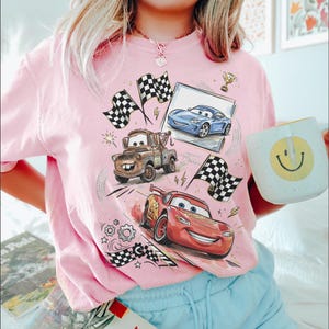 Op de afbeelding: Lichtroze T-shirt met een afbeelding van geanimeerde raceauto's, geblokte vlaggen en een trofee. Het ontwerp bevat afbeeldingen van een blauwe auto, een bruine takelwagen en een rode raceauto. Een witte mok met een gele smiley wordt in de afbeelding vastgehouden.