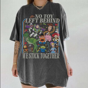 May include: Dark gray t-shirt featuring a colorful graphic of animated characters with the text "NO TOY LEFT BEHIND" and "WE STICK TOGETHER". The design includes a dinosaur, space ranger, cowboy, and other characters.