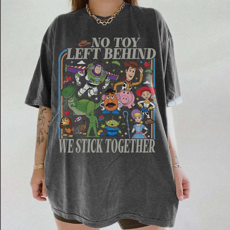 May include: Dark gray t-shirt featuring a colorful graphic of animated characters with the text "NO TOY LEFT BEHIND" and "WE STICK TOGETHER". The design includes a dinosaur, space ranger, cowboy, and other characters.