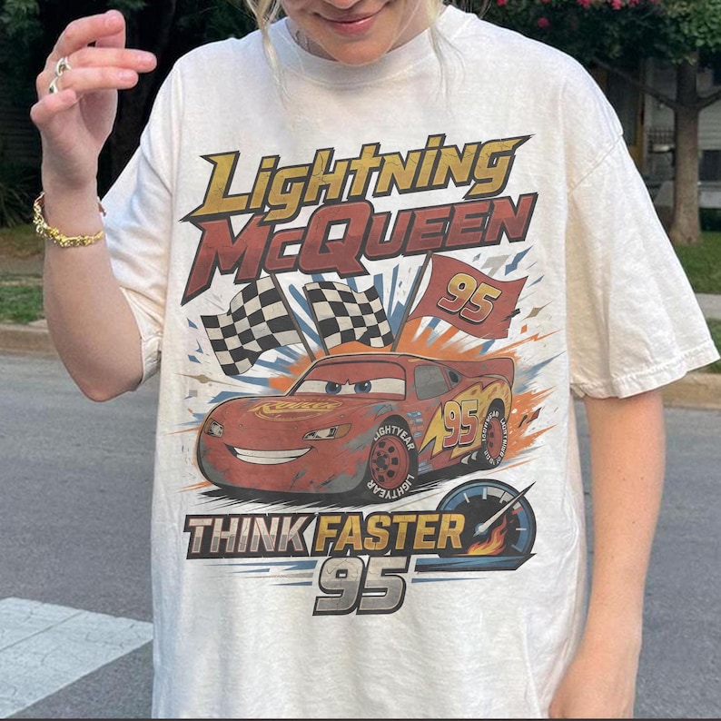 K&ouml;nnte beinhalten: Cremefarbenes T-Shirt mit einer Grafik von Lightning McQueen aus dem Film Cars. Das Design enth&auml;lt den Text "Lightning McQueen" und "Think Faster 95" mit Schachflaggen und einem Tachometer.