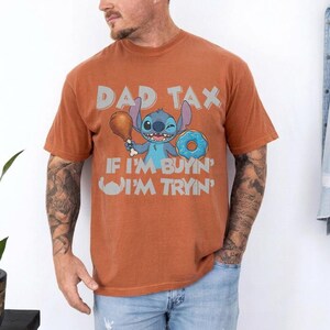 May include: Rust-colored t-shirt featuring a cartoon Stitch character holding a drumstick and a donut. The shirt has the text "DAD TAX" above Stitch and "IF I'M BUYIN' I'M TRYIN'" below. The shirt is made of a soft material.