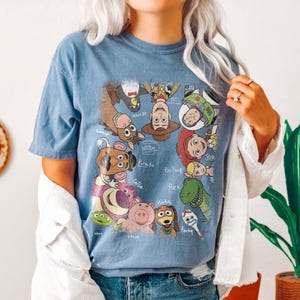 May include: Blue t-shirt featuring a graphic of various characters from the animated film Toy Story. The characters include Woody, Buzz Lightyear, Jessie, and others. The shirt is a light blue color.