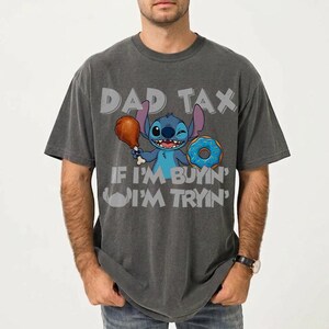 May include: Dark gray t-shirt featuring the character Stitch from Lilo & Stitch. Stitch is holding a drumstick and a donut. The shirt has the text "DAD TAX" and "IF I'M BUYIN' I'M TRYIN'".